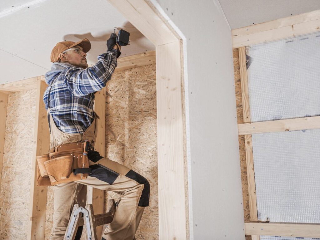 A general contractor installing drywall inside a partially constructed home, wearing a tool belt and safety gear.